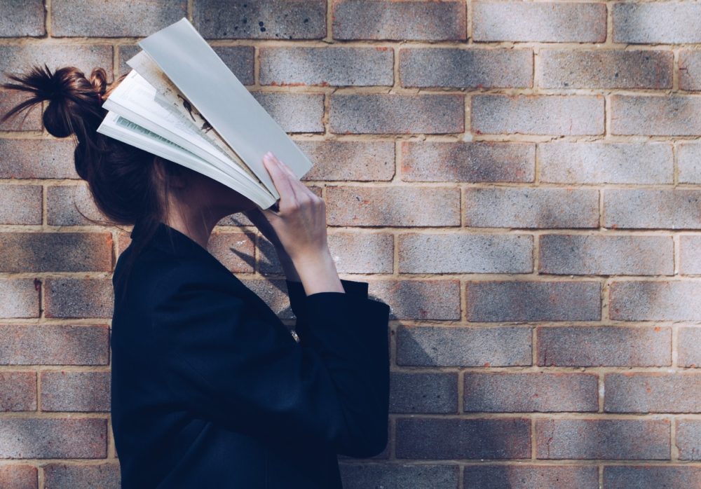 A woman holding a book over her face and standing beside a brick wall