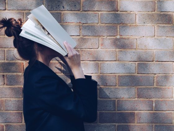 A woman holding a book over her face and standing beside a brick wall